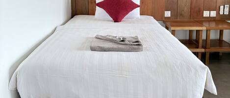 A neatly made bed with a white comforter and a red accent pillow, a wooden headboard, a bedside table, and a wall-mounted lamp.