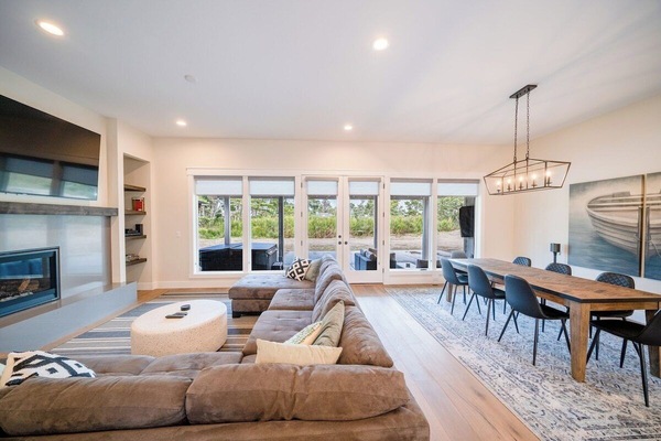 Relax in a spacious open-concept living area with a plush sectional, 82” TV, cozy fireplace, and backyard views—perfect for unforgettable gatherings!