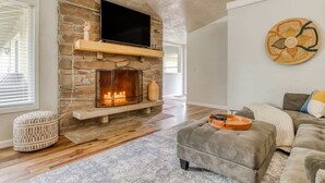 Una sala de estar con chimenea de piedra, un televisor de pantalla plana montado encima y un sofá gris con un tapete estampado.