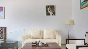 Una sala de estar moderna con un sofá color beige, una mesa de centro de madera y una mesa de vidrio. Se observa un cuadro en la pared y una lámpara en el suelo.