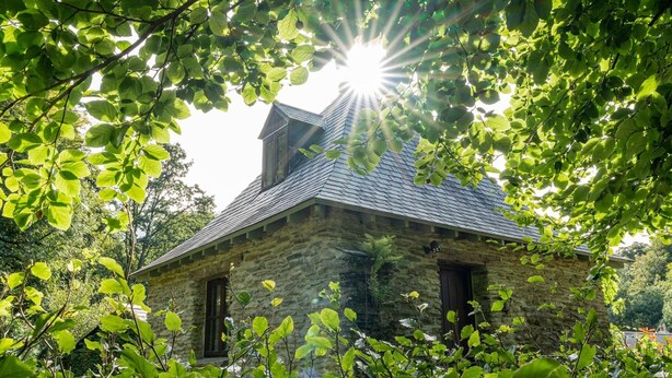 A stone house with a sloped roof, surrounded by lush green foliage.