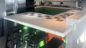 Eine Küche mit grünen Schränken, einem Edelstahlkühlschrank, einer großen Kochinsel mit Hockern und Pendelleuchten.