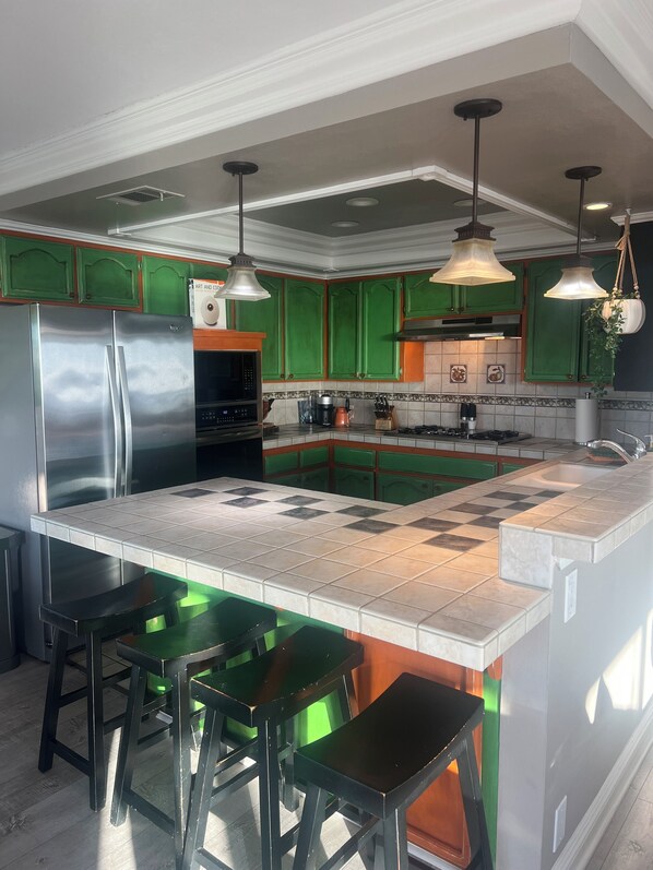 Une cuisine avec des meubles de couleur verte, un réfrigérateur en acier inoxydable, un grand îlot central avec des tabourets et des luminaires suspendus.