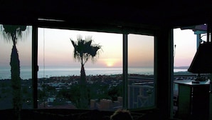 Ein Zimmer mit großem Fenster, das Blick auf einen Sonnenuntergang, Palmen und eine ferne Skyline bietet.