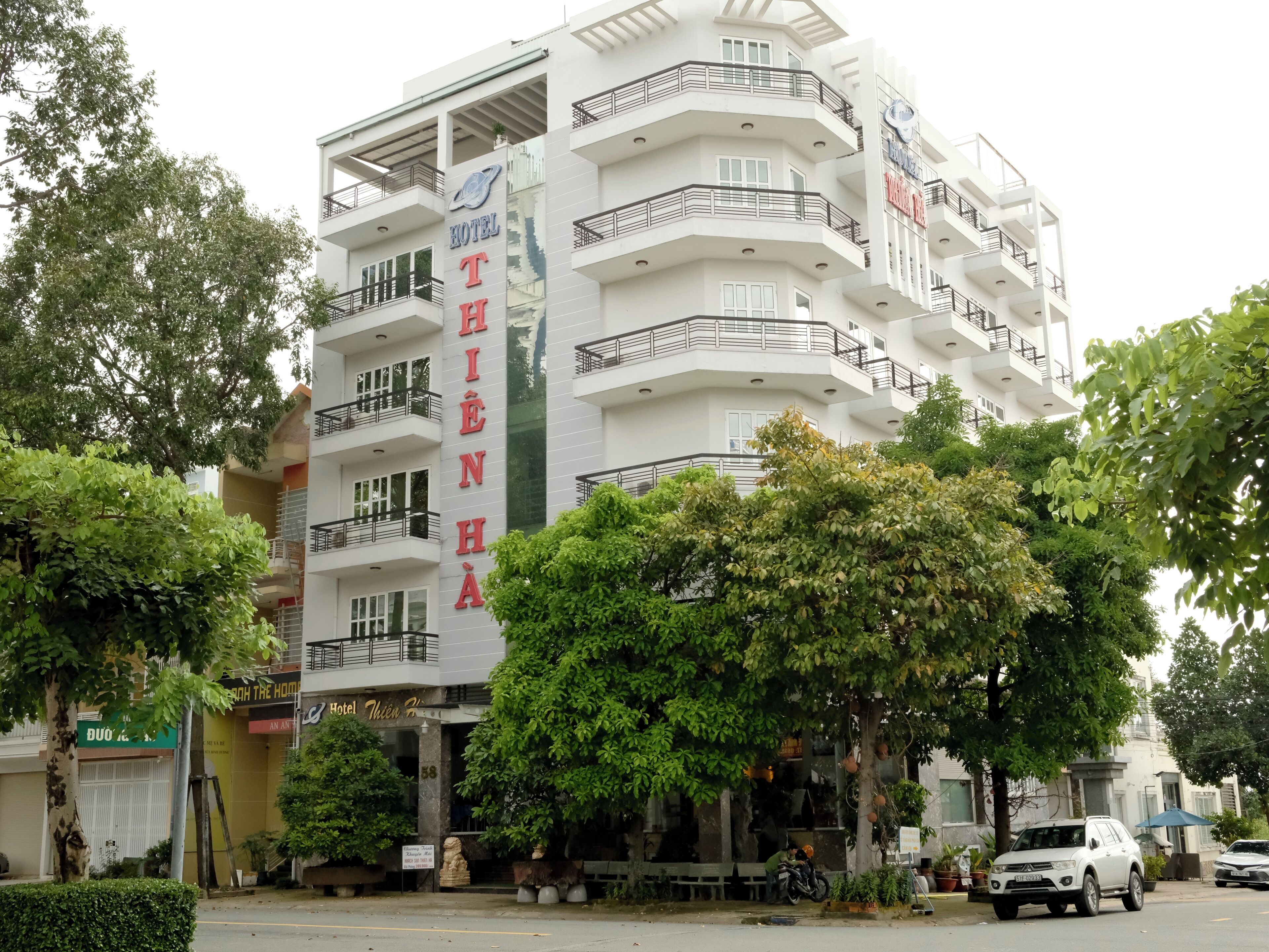 A multi-story hotel with balconies, a sign indicating it is a hotel, and a street view with parked cars and trees.