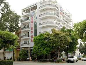 Ein mehrstöckiges Hotel mit Balkonen, einem Hinweisschild, das es als Hotel kennzeichnet, und eine Straßenszene mit parkenden Autos und Bäumen.