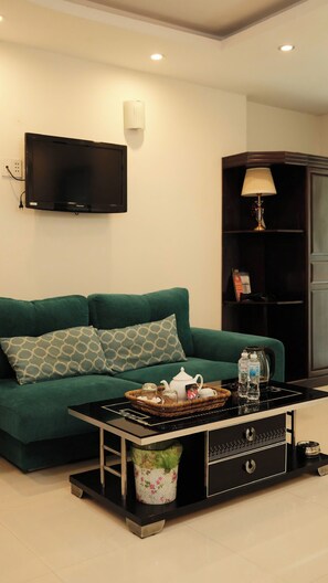 Ein Wohnzimmer mit einem grünen Sofa, einem Flachbildfernseher, einem Couchtisch mit Tablett, einem blumigen Mülleimer und einem Bücherregal.