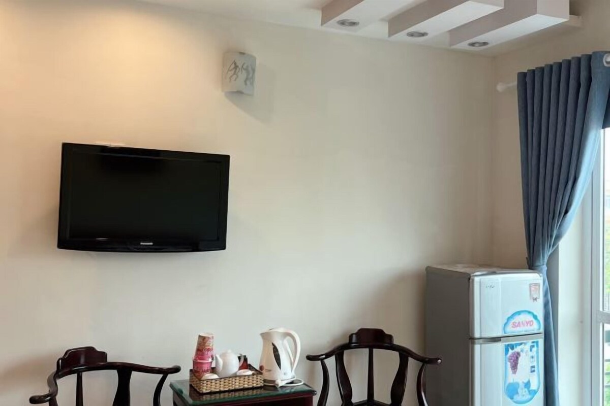 Ein Zimmer mit einem an der Wand befestigten Flachbildfernseher, einem kleinen Tisch mit Wasserkocher und Tassen, zwei Holzstühlen und einem Kühlschrank mit Sanyo-Logo.