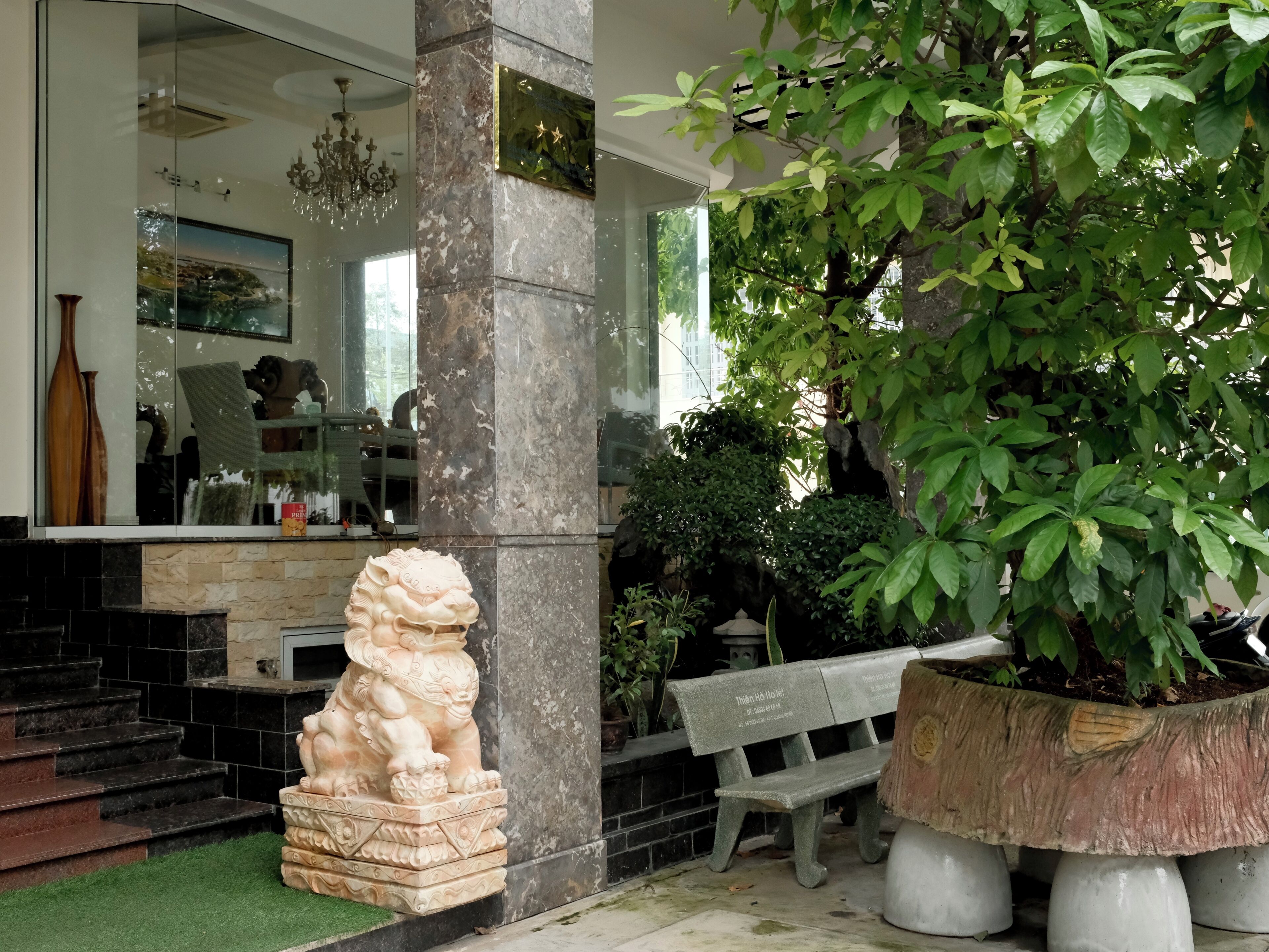 A stone lion statue, a bench, and a large potted plant in a courtyard with a marble column and a glass wall reflecting interior furnishings.