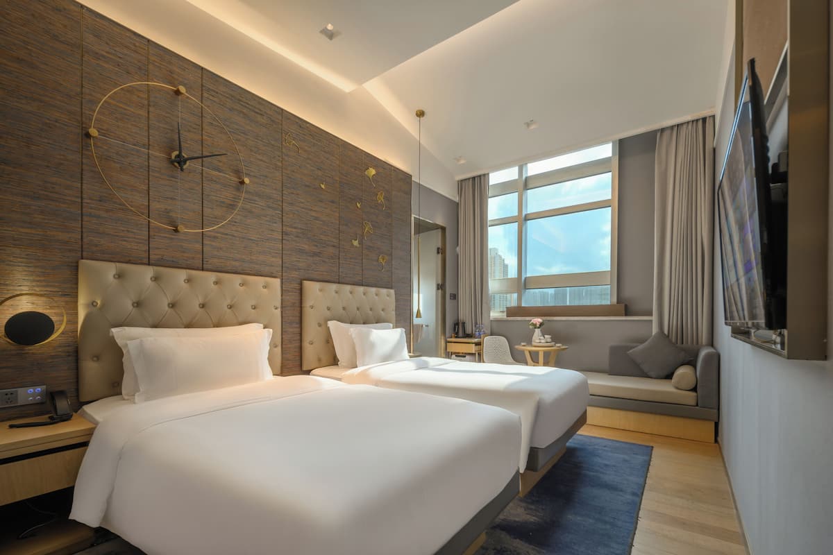 A modern hotel room with two large beds, a seating area, a large window, and a wall-mounted clock.