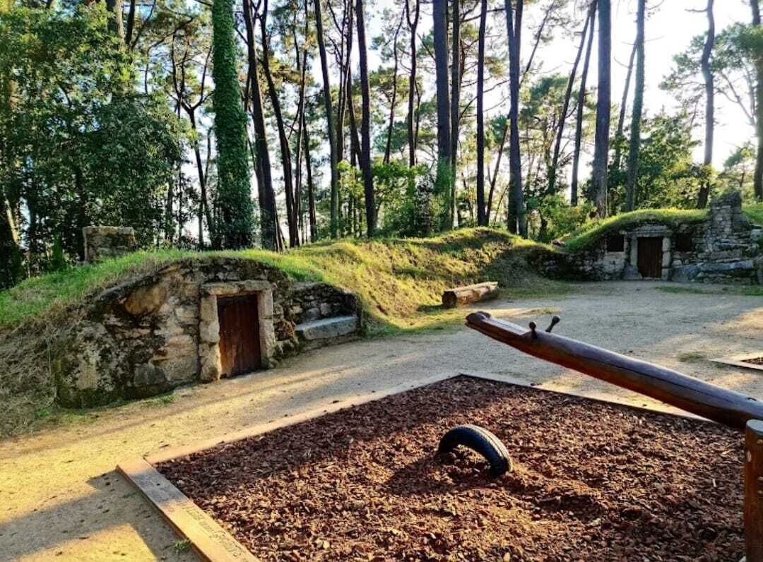 Een park met een pad van aarde, een stenen constructie met een deur en een speelplaats met een wipkippen en een band.