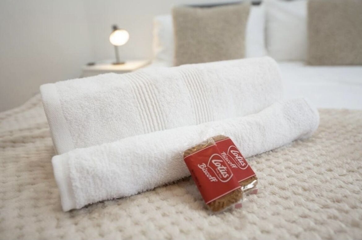 A neatly folded white towel and a small box of Lotus Biscotti on a textured beige bedspread.