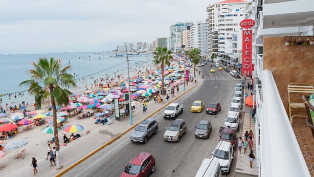 Un hotel frente a la playa con vista al océano, una calle bordeada de coches aparcados y sombrillas de colores.