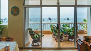 Una habitación con puerta de vidrio corredera que da a un balcón con vista al mar y barcos. Hay sillas de mimbre y una mesa en el balcón.
