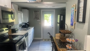 A compact kitchen with white cabinets, a black stove, and a microwave. There are various kitchen items on the counter, including a coffee maker and a toaster. A door with a 'No Smoking' sign is visible, and a window allows natural light into the room. - Mango Beach Retreat (Lake Worth)