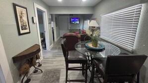 A dining area with a glass table, chairs, a vase with flowers, and a television in the background. - Mango Beach Retreat (Lake Worth)
