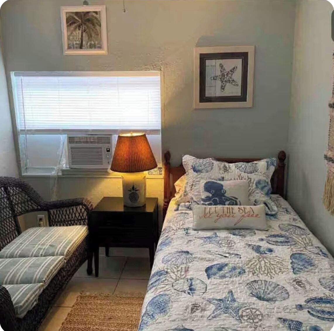 Ein Schlafzimmer mit Bett, Nachttisch, Stuhl, Fenster mit Jalousien und Wandbildern.