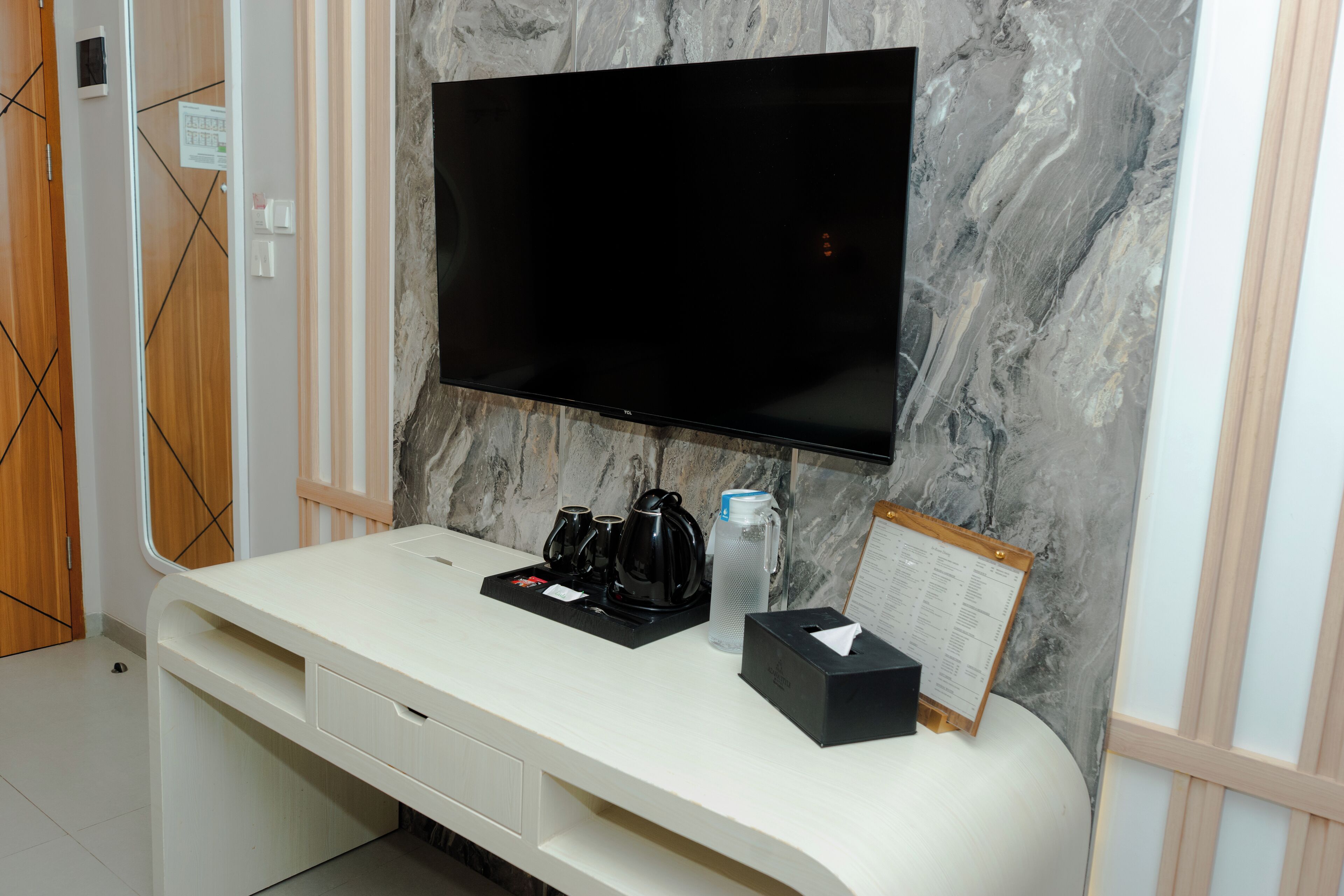 A hotel room with a flat-screen TV mounted on a marble wall, a white console table with a coffee maker, a water bottle, and a tissue box.