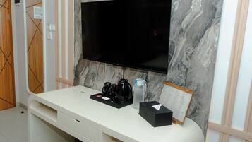 Habitación de hotel con una televisión de pantalla plana montada en una pared de mármol, una mesa blanca con mesita de noche que incluye una cafetera, una botella de agua y una caja de pañuelos.