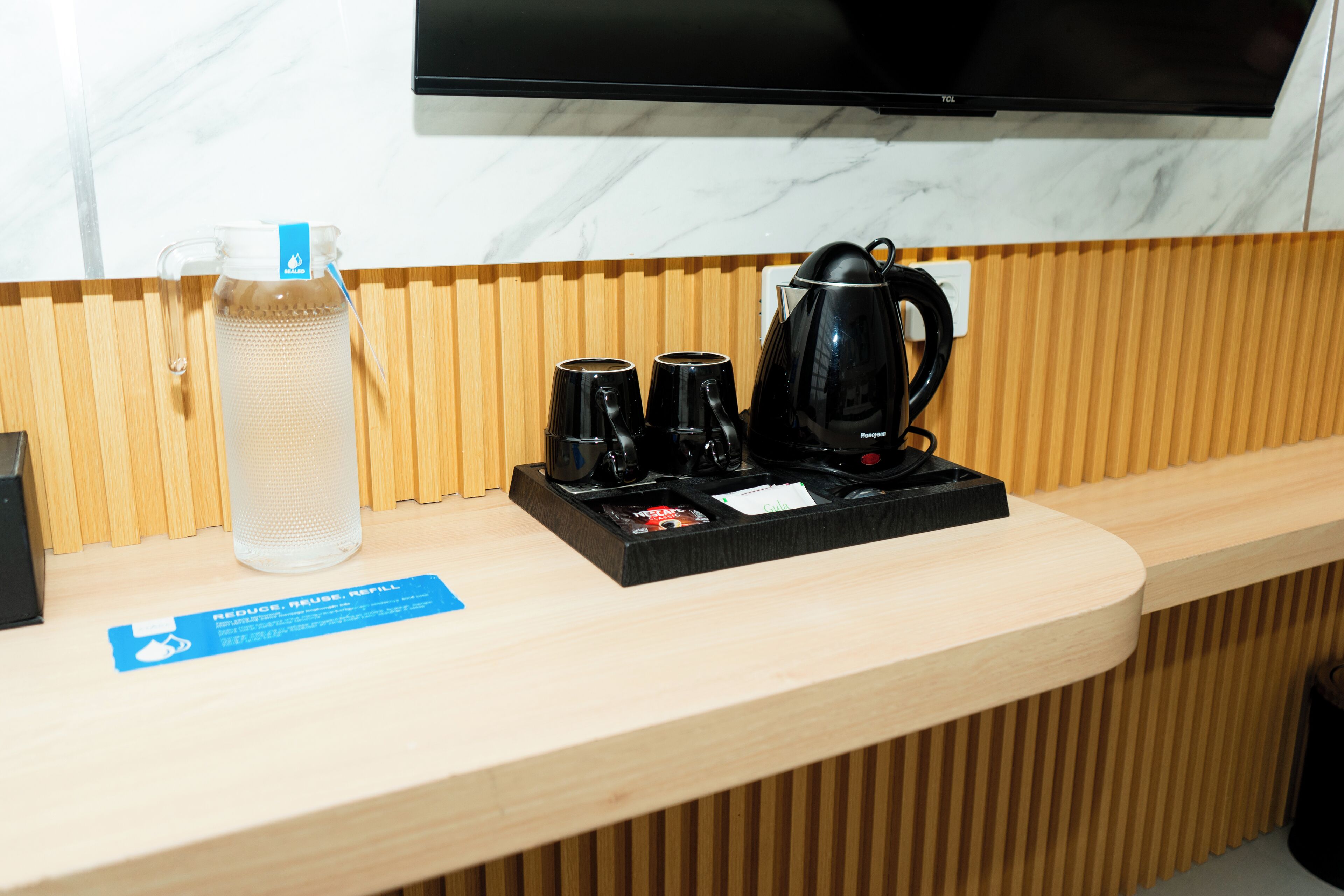 A hotel reception area with a marble wall, wooden paneling, a black coffee maker, two black cups, a glass water bottle, and a sign encouraging guests to reduce, reuse, and refill.