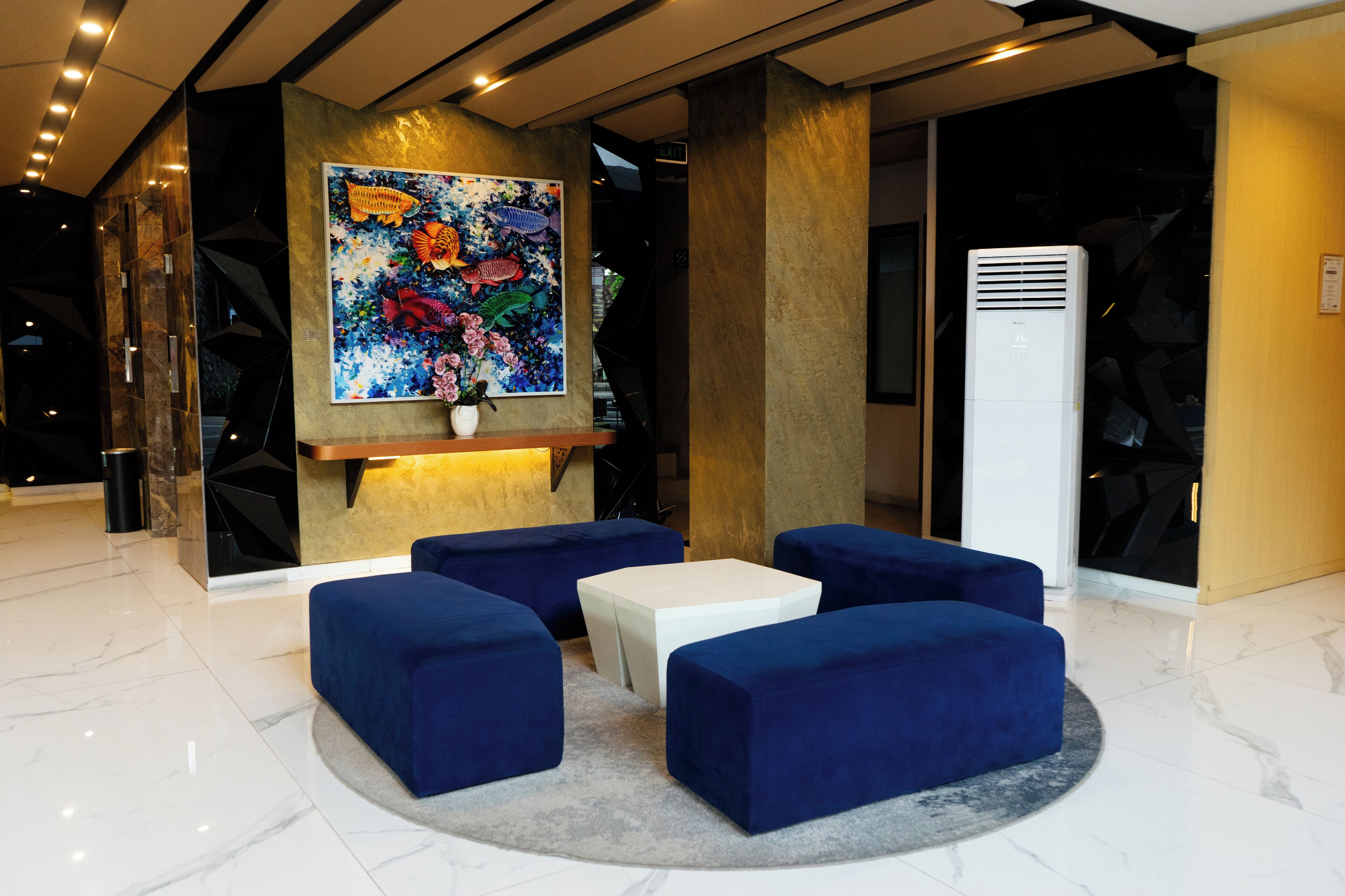 A lobby with a large colorful fish painting, blue cushioned seating, a white coffee table, and a modern air conditioning unit.