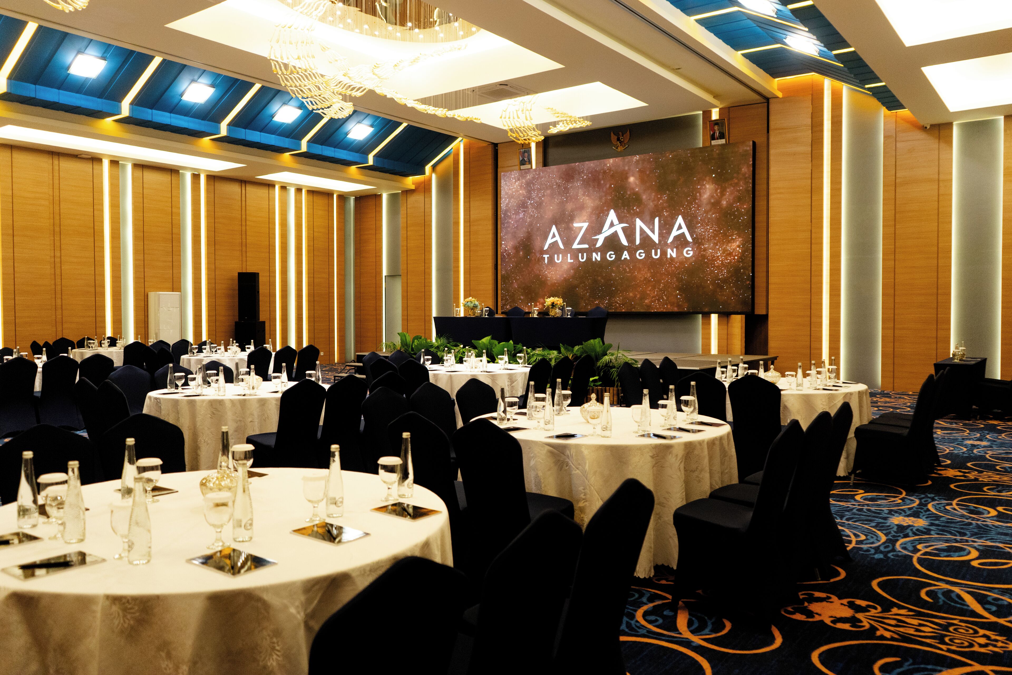 A conference room with round tables, black chairs, and a large screen displaying "AZANA TULUNGAGUNG".