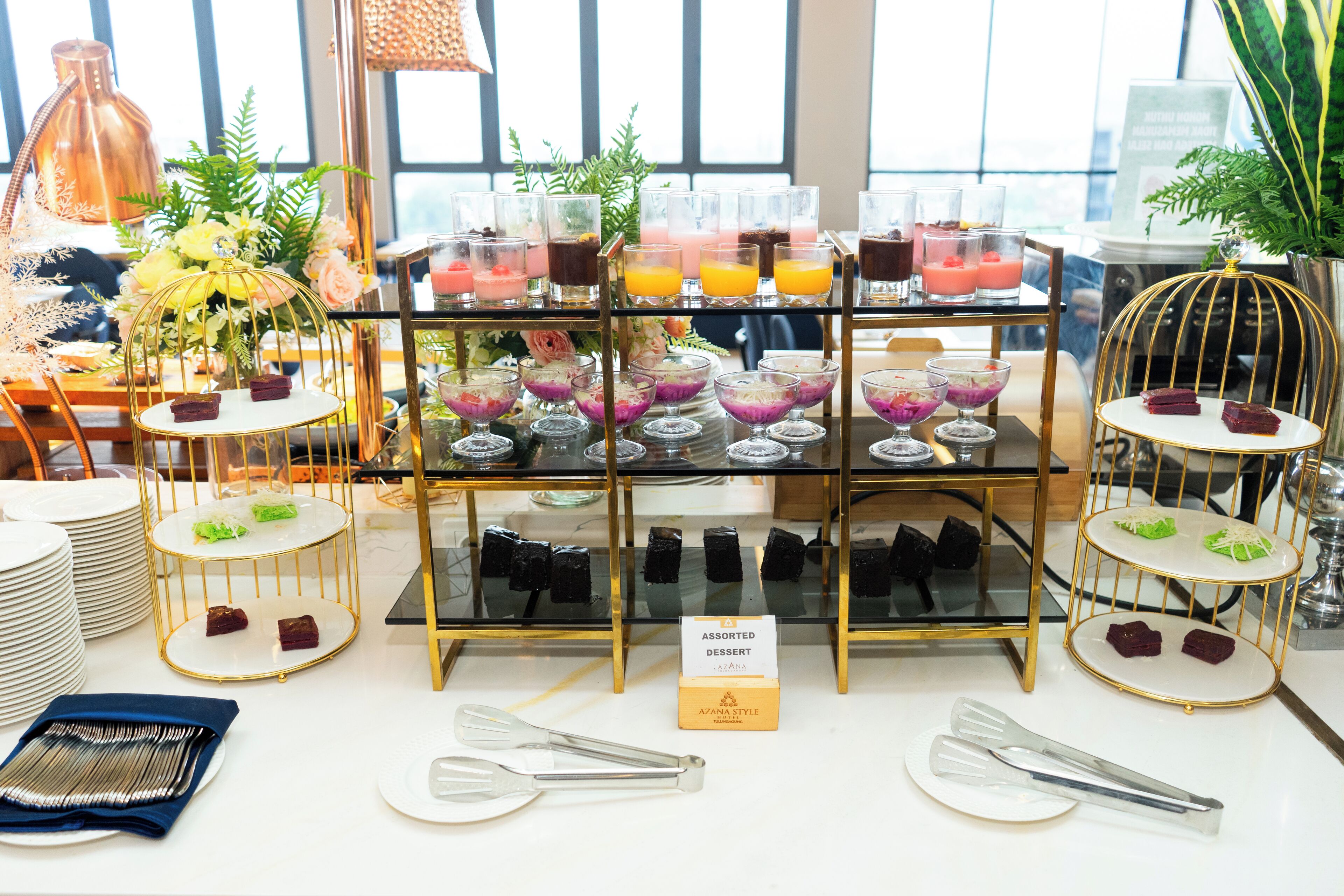 Una exhibición de postres con variedad de dulces y bebidas, un letrero que dice 'Postres Variados' y una jaula de pájaros con adornos.