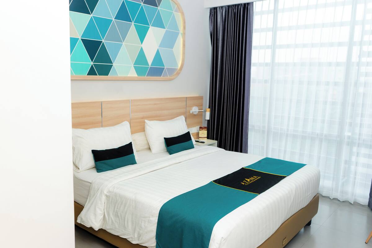 A hotel room with a large bed, a wooden headboard, a geometric wall art piece, a nightstand with a lamp, and a window with curtains.
