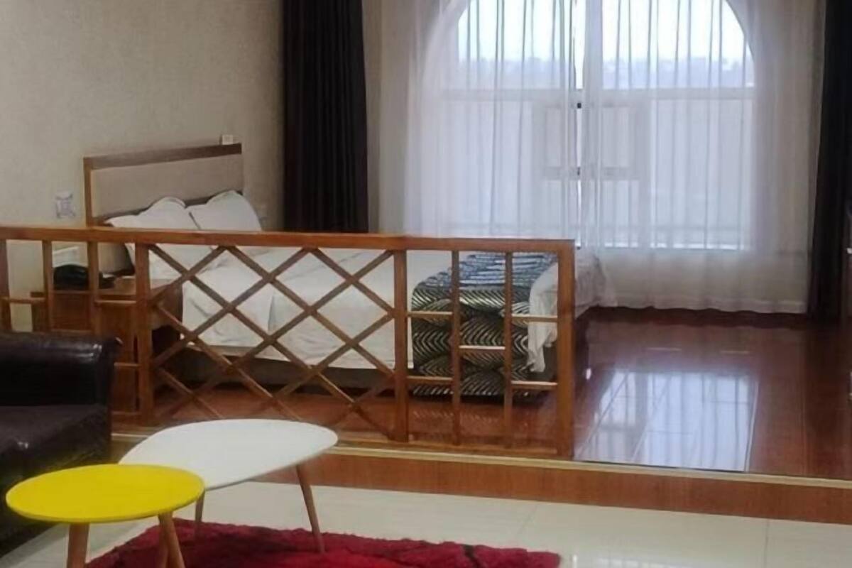 Una habitación con cama, un separador de madera, un sofá, una pequeña mesa redonda y un ventanal con cortinas translúcidas.
