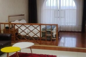 Een kamer met een bed, een houten scheidingswand, een bank, een kleine ronde tafel en een groot raam met dunne gordijnen.
