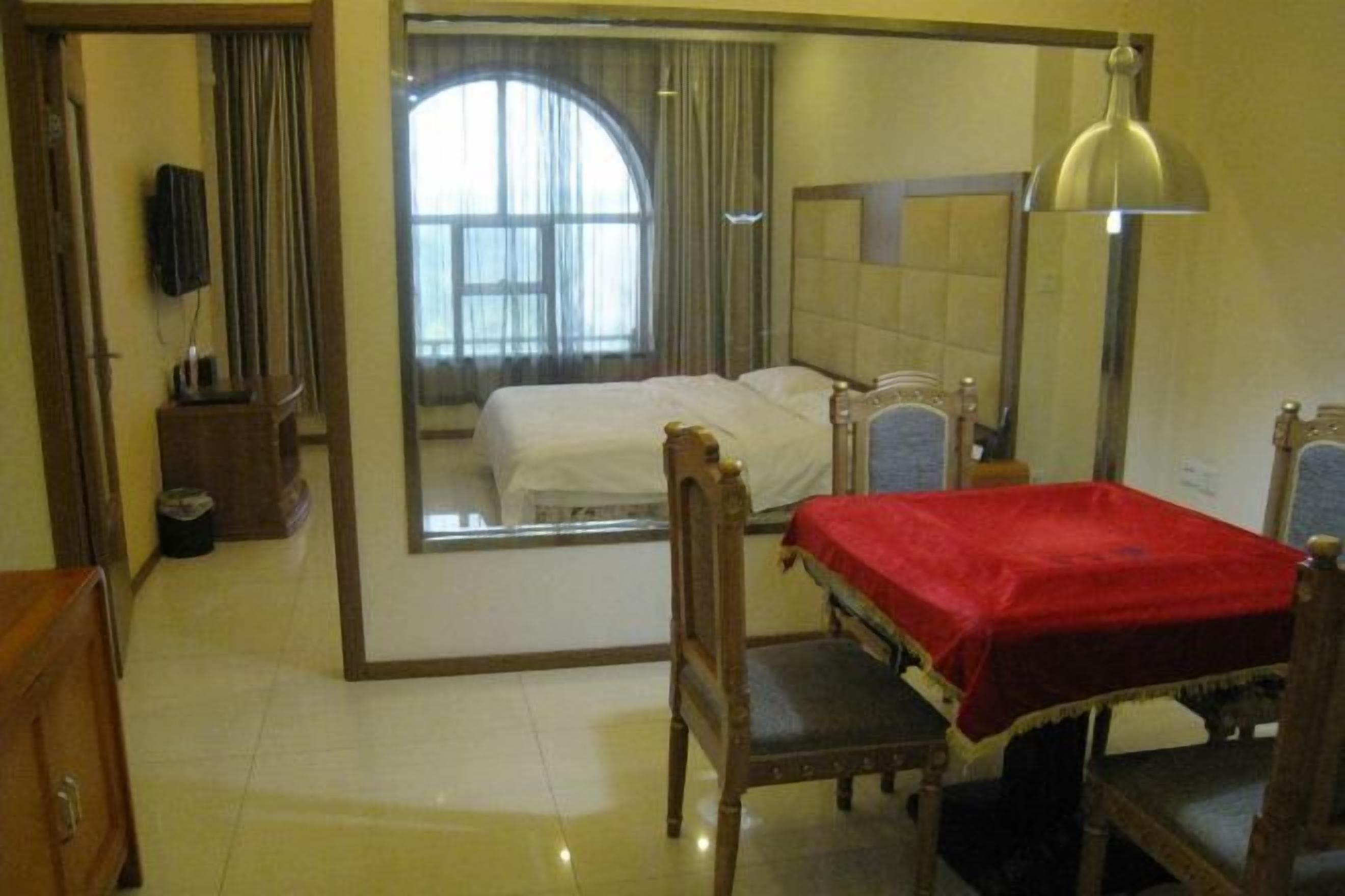A hotel room with a large mirror, a bed with a red blanket, a wooden chair, a desk with a lamp, and a view of a window with curtains.