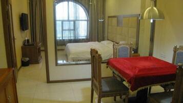 A hotel room with a large mirror, a bed with a red blanket, a wooden chair, a desk with a lamp, and a view of a window with curtains.