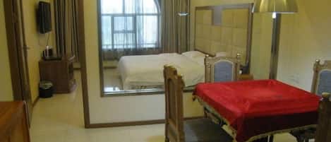 A hotel room with a large mirror, a bed with a red blanket, a wooden chair, a desk with a lamp, and a view of a window with curtains.