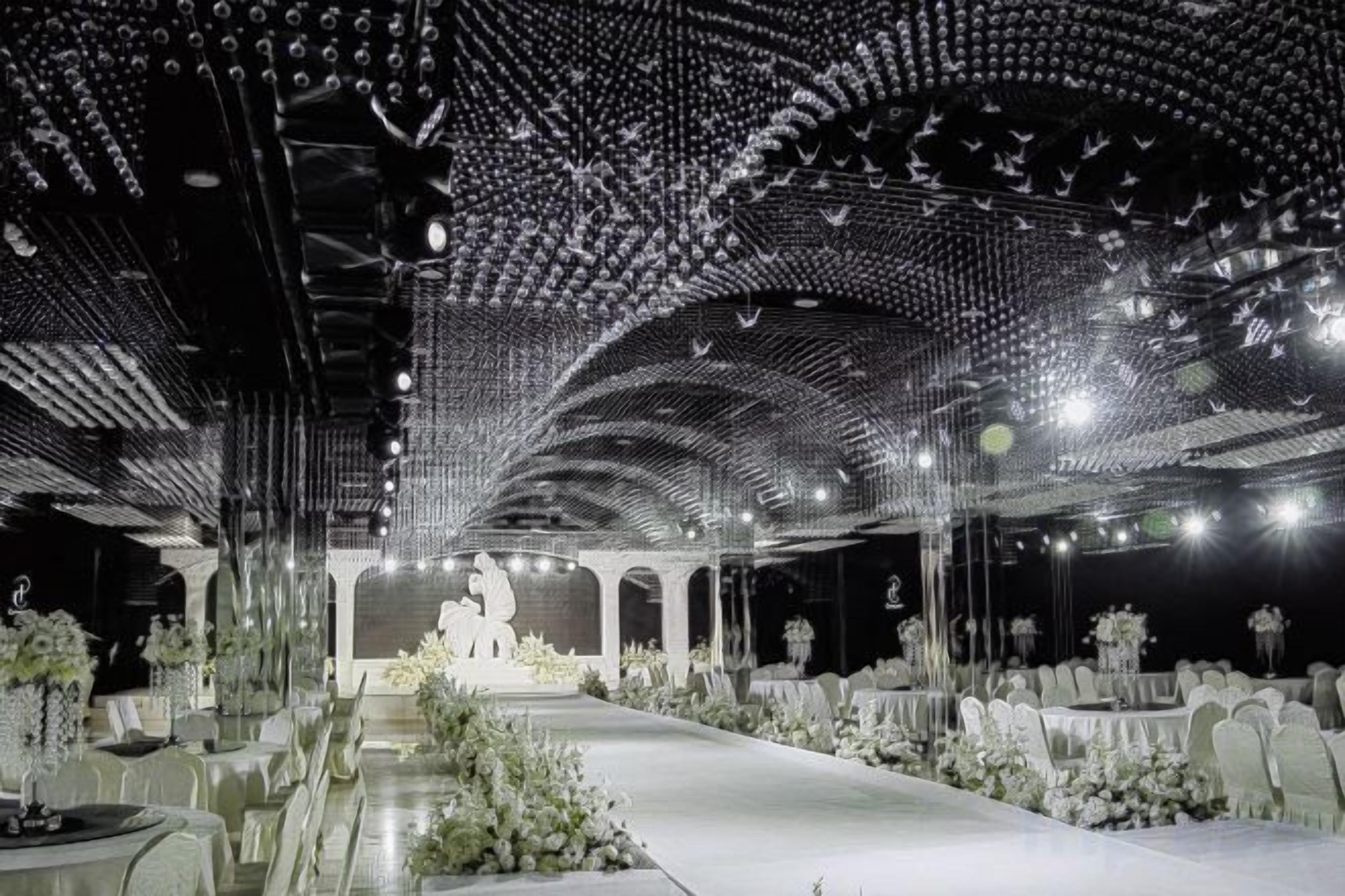 An indoor venue with a long table set for a formal event, a central stage, and a ceiling with a geometric pattern.