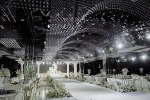 An indoor venue with a long table set for a formal event, a central stage, and a ceiling with a geometric pattern.