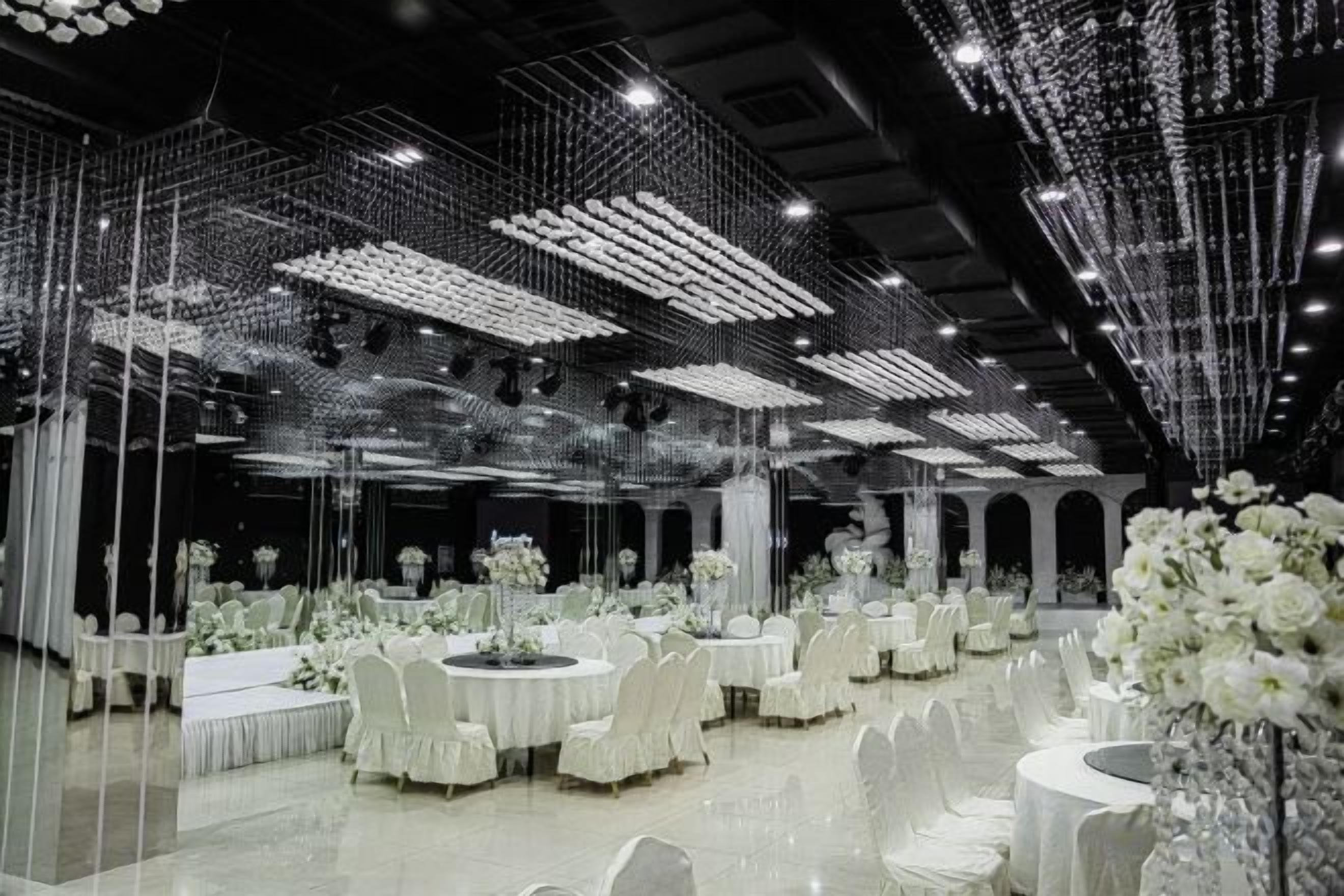A banquet hall with round tables, white floral centerpieces, and a ceiling adorned with hanging lights.
