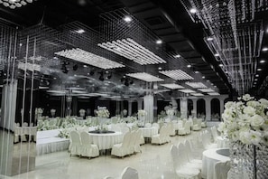 Un salón de banquetes con mesas redondas, centros de mesa con flores blancas y un techo decorado con luces colgantes.