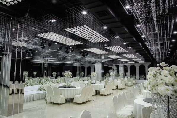 A banquet hall with round tables, white floral centerpieces, and a ceiling adorned with hanging lights.