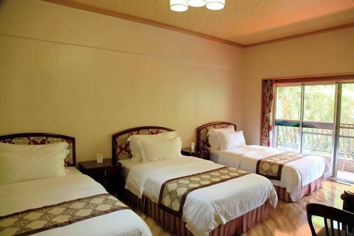 A hotel room with three beds, a wooden floor, and a large window with a view of greenery.