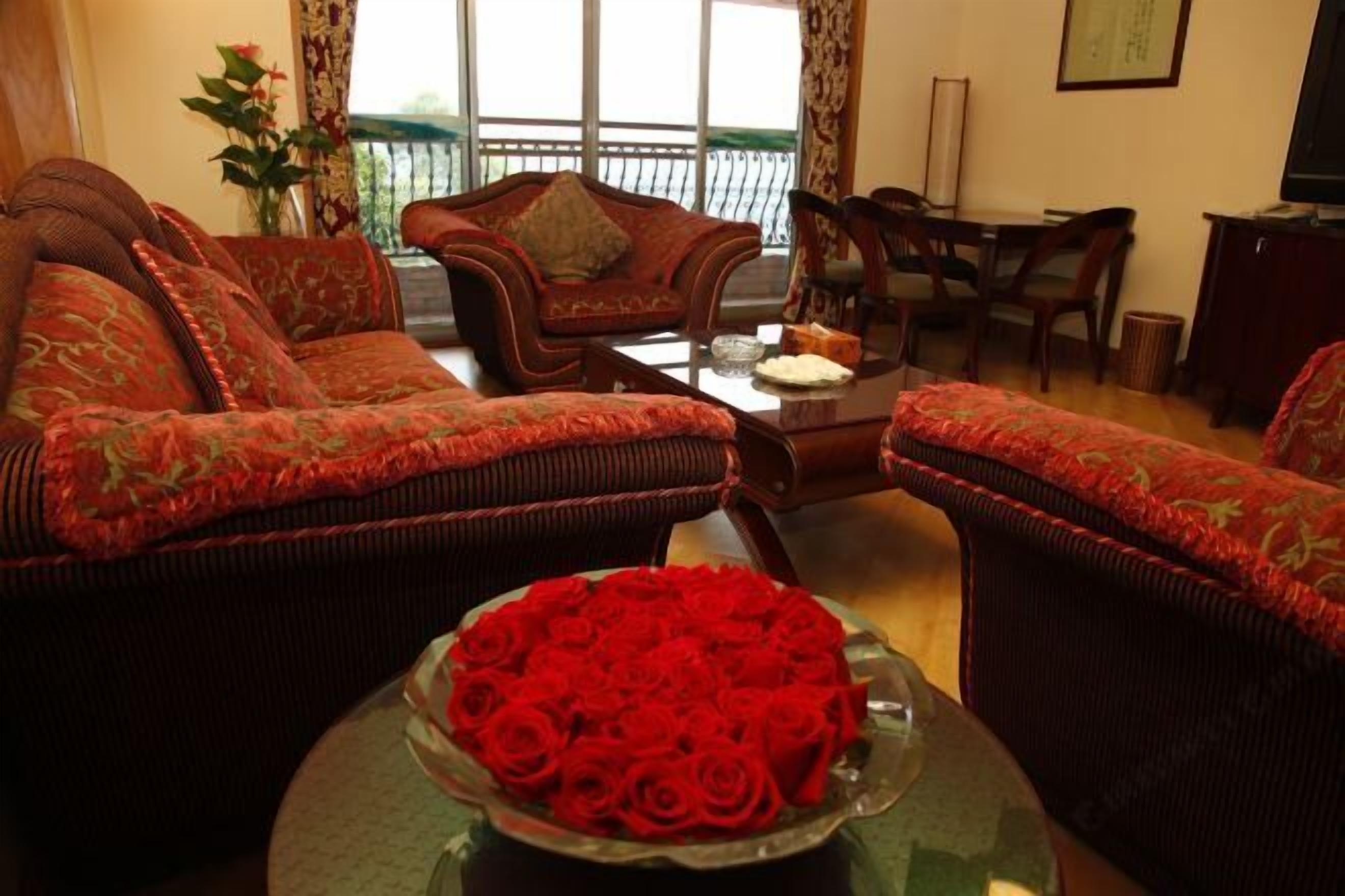 Un salon avec deux canapés rouges, un guéridon surmonté d’un vase en verre rempli de roses rouges, et un coin repas avec des meubles en bois.