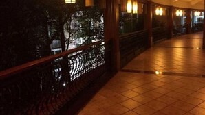 Corredor com piso em mosaico e guarda-corpos de madeira, com luminárias fixadas na parede e vista para árvores do lado de fora.