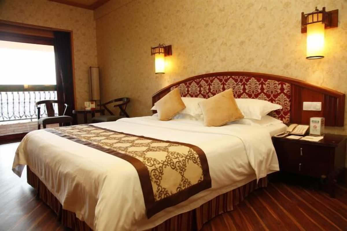 A hotel room with a large bed, a wooden headboard, two bedside lamps, a nightstand with a phone, and a window with a view.