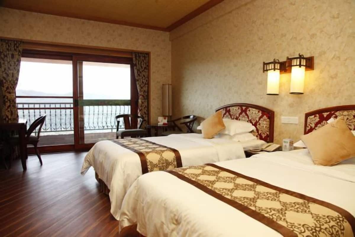 A hotel room with two beds, a desk, and a chair. There is a balcony with a railing and a view of the sea.