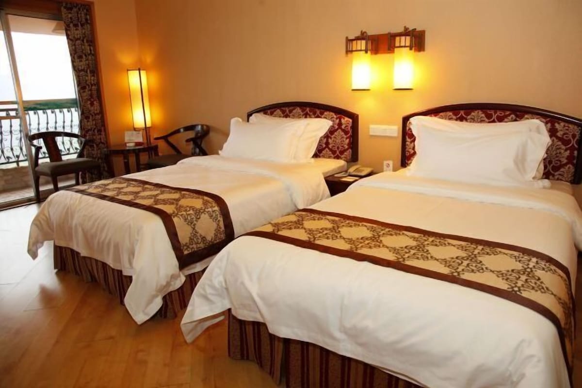 A hotel room with two beds, a chair, a nightstand, a lamp, and a view of the outside.