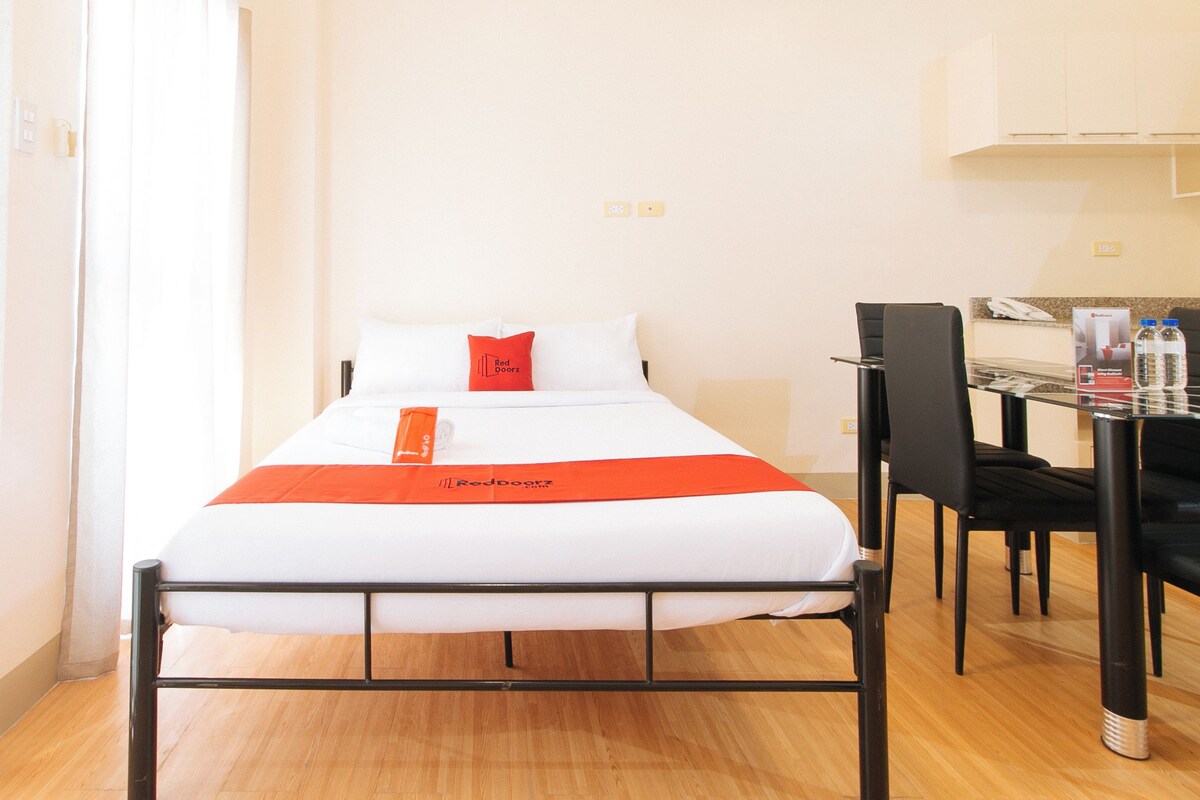 Una cama individual con colchón blanco y colcha naranja, un cojín rojo y una pequeña mesa con sillas.