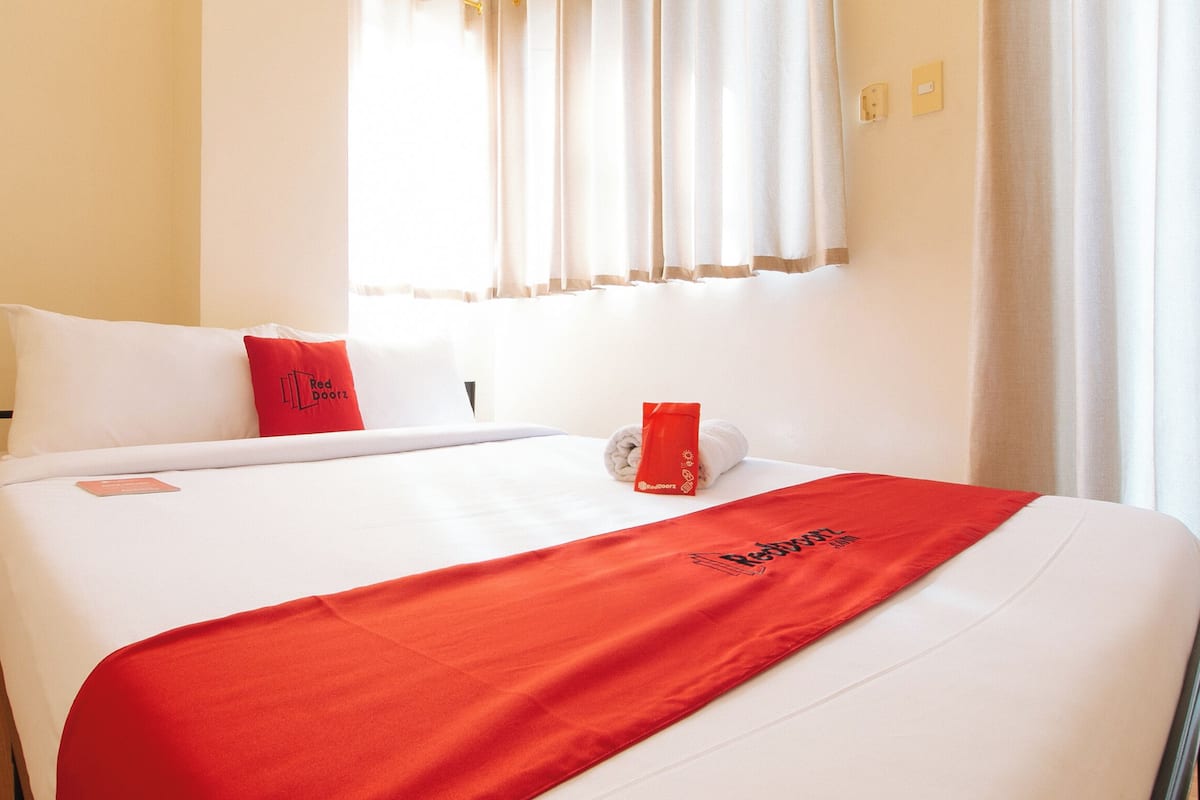 Una habitación de hotel con cama, ropa de cama blanca, una colcha roja, almohada con logotipo y ventana con cortinas.
