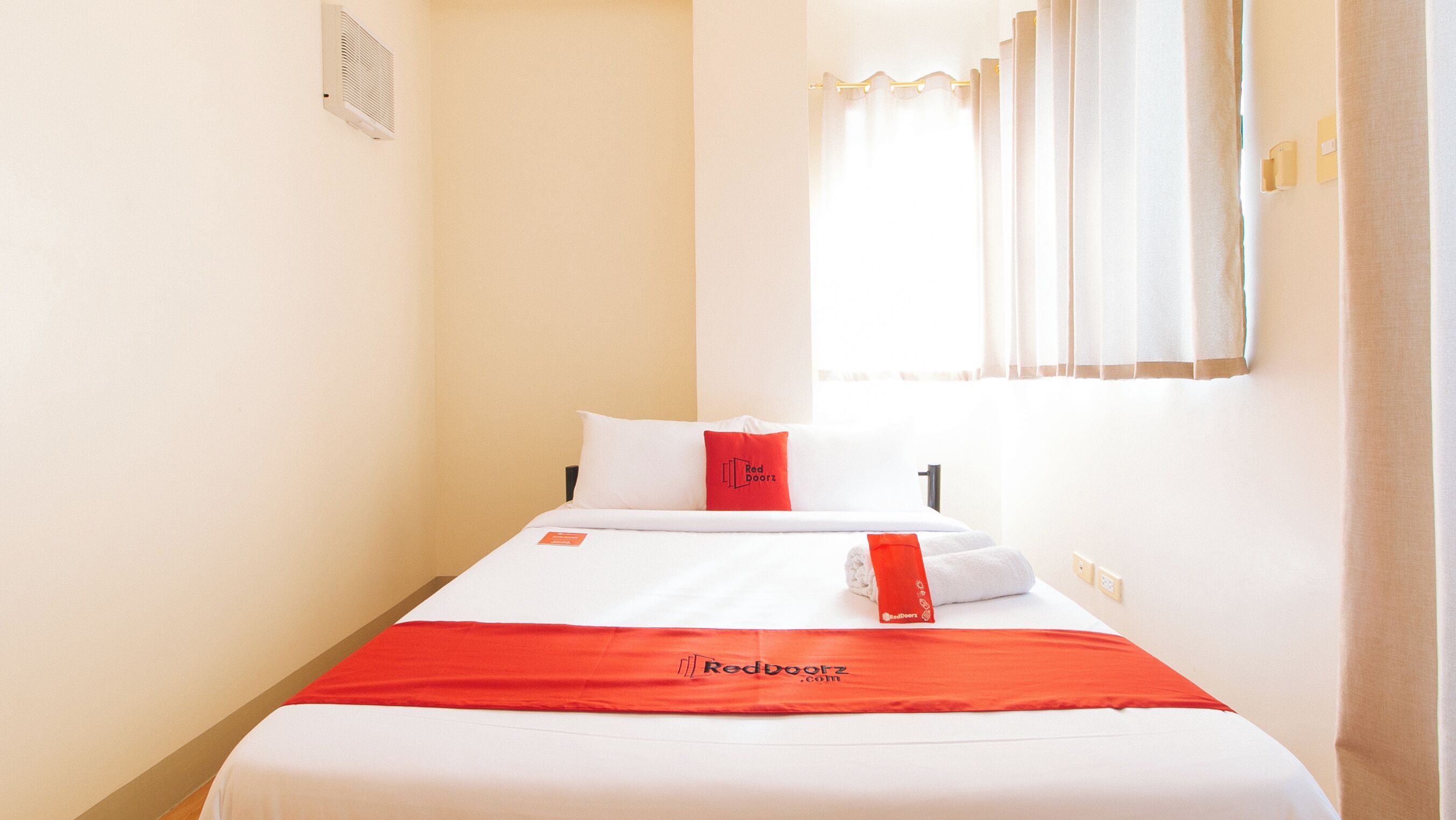 A hotel room with a bed, a red blanket, a pillow, a window with curtains, and a wall-mounted air vent.