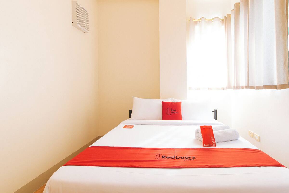 Habitación de hotel con cama, manta roja, almohada, ventana con cortinas y un respiradero en la pared.