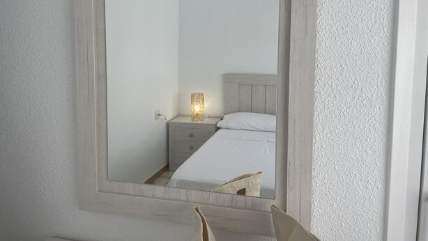 Un miroir avec un luminaire sur une table de chevet, un lit avec une literie blanche et une tête de lit en bois.