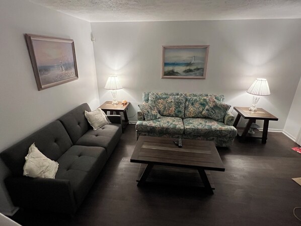 A living room with a dark grey sofa, a floral-patterned armchair, a wooden coffee table, and two side tables with lamps. There are two framed paintings on the walls.